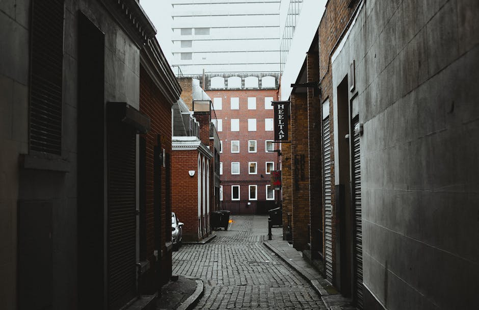 A narrow urban alleyway filled with a large, wheeled waste container in the foreground, covered with a heavy, weathered fabric sack, situated on a cracked pavement with dark stains and debris scattere