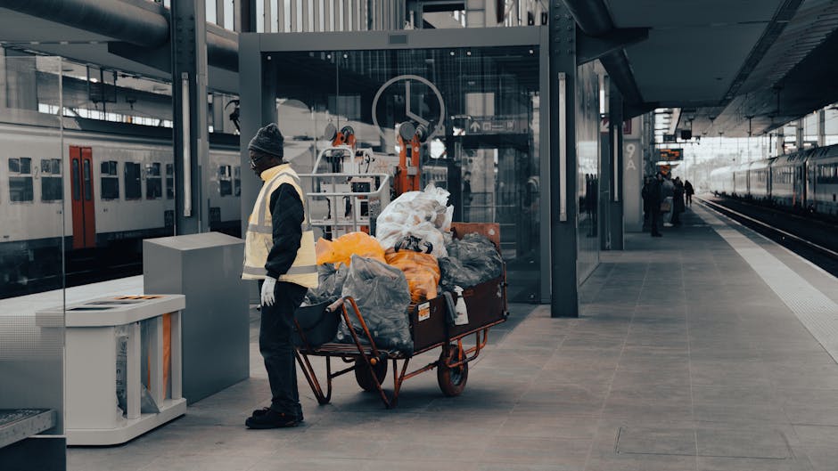 A person wearing a dark jacket, grey beanie, and white gloves stands on a modern train station platform beside a large metal-wheeled trolley loaded with miscellaneous waste and rubbish bags of various